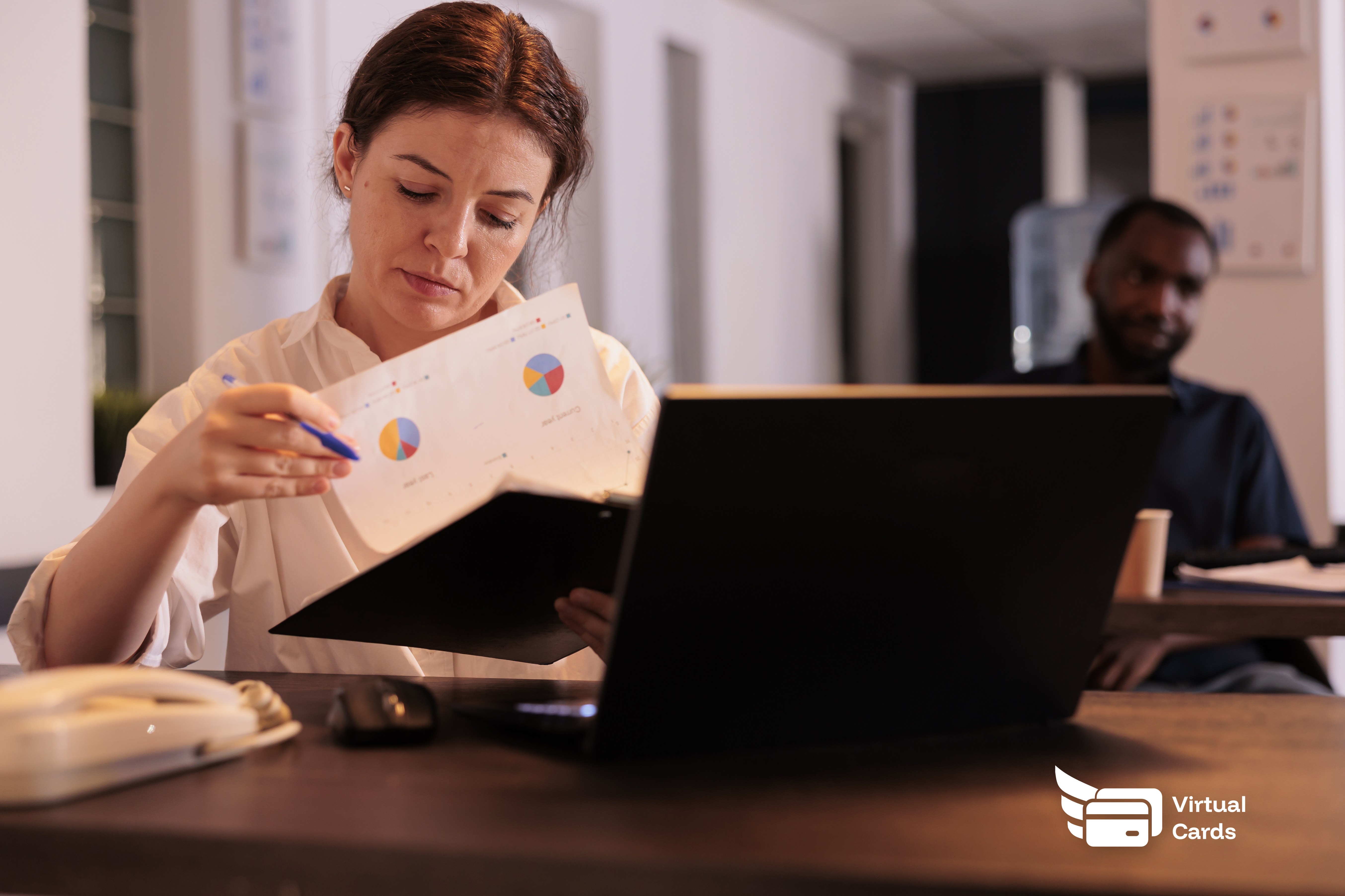 A project manager at a desk reviewing a laptop with project budgets and spending reports, a virtual Visa card visible on screen.